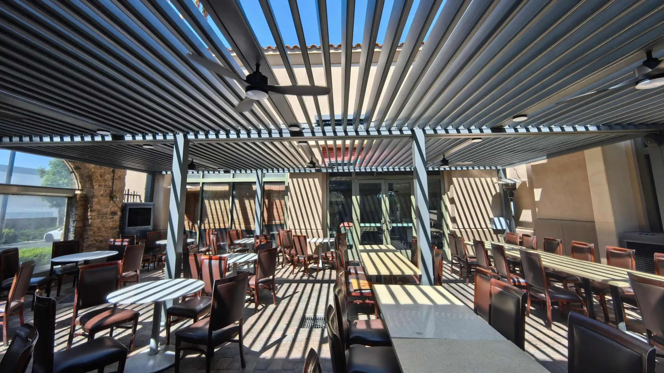 Sunlit restaurant outdoor dining area with louvered roof, warm sunlight streaming through slats over tables