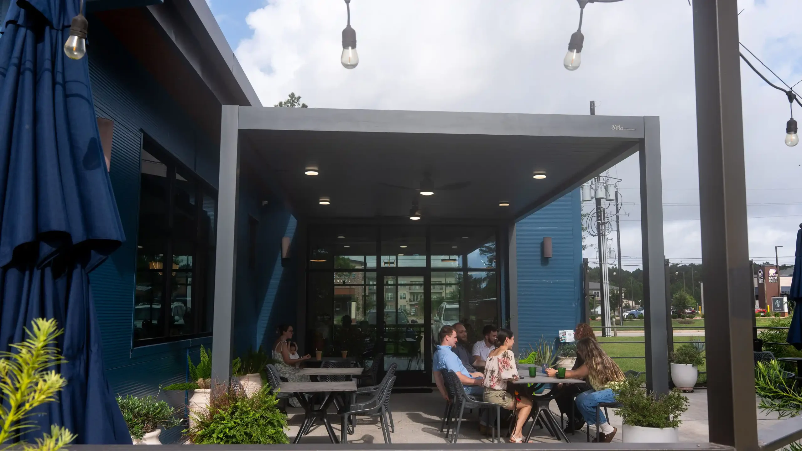 exterior shot cafe patio covered by dark framed solar pergola customers dining blue building background